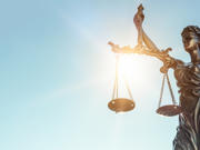 A bronze statue of Lady Justice holding balanced scales in her left hand and a sword in her right, with sunlight shining through the scales against a clear blue sky.