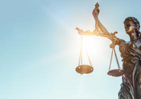 A bronze statue of Lady Justice holding balanced scales in her left hand and a sword in her right, with sunlight shining through the scales against a clear blue sky.