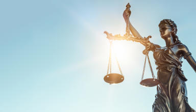 A bronze statue of Lady Justice holding balanced scales in her left hand and a sword in her right, with sunlight shining through the scales against a clear blue sky.