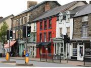 A high street in Wales. There is a pedestrian crossing across a road in the middle of the image, and behind that there are businesses and shops in  buildings of different colours.