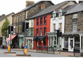 A high street in Wales. There is a pedestrian crossing across a road in the middle of the image, and behind that there are businesses and shops in  buildings of different colours.
