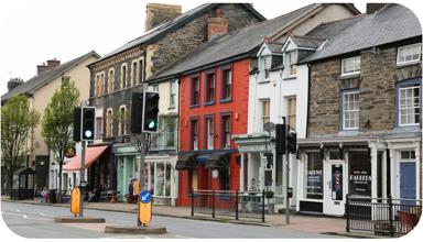 A high street in Wales. There is a pedestrian crossing across a road in the middle of the image, and behind that there are businesses and shops in  buildings of different colours.