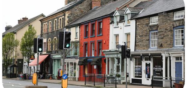 A high street in Wales. There is a pedestrian crossing across a road in the middle of the image, and behind that there are businesses and shops in  buildings of different colours.