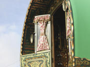 Close-up view of a traditional, ornately decorated caravan with a green exterior. The image focuses on an open window with floral-patterned curtains tied back, revealing part of the interior. The caravan features intricate gold and red designs on the wood.