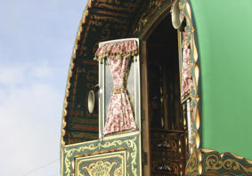 Close-up view of a traditional, ornately decorated caravan with a green exterior. The image focuses on an open window with floral-patterned curtains tied back, revealing part of the interior. The caravan features intricate gold and red designs on the wood.