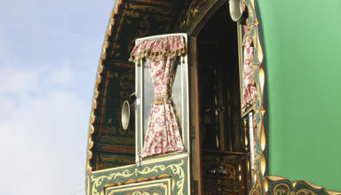 Close-up view of a traditional, ornately decorated caravan with a green exterior. The image focuses on an open window with floral-patterned curtains tied back, revealing part of the interior. The caravan features intricate gold and red designs on the wood.