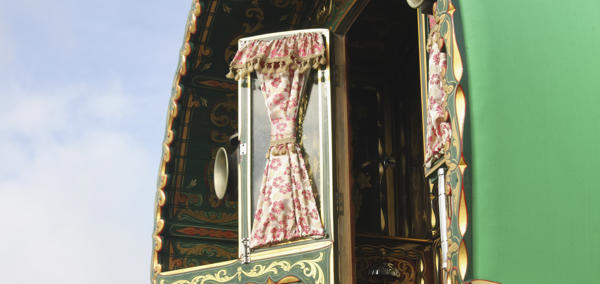 Close-up view of a traditional, ornately decorated caravan with a green exterior. The image focuses on an open window with floral-patterned curtains tied back, revealing part of the interior. The caravan features intricate gold and red designs on the wood.