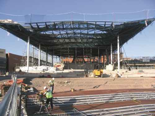 Images shows two construction workers wearing hi-visibility vests and hard hats operating machinery in the foreground. Behind them is the steel framework of the Senedd building below a blue sky.