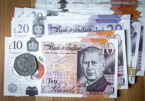 Ten pound sterling note on top of other notes, a 50 pence piece is on top of the ten pound note. The note and coin carry the image of the King.