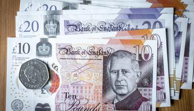 Ten pound sterling note on top of other notes, a 50 pence piece is on top of the ten pound note. The note and coin carry the image of the King.