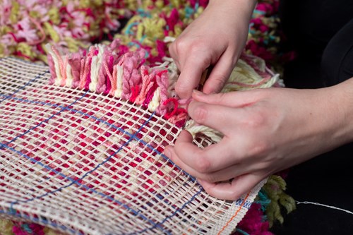 A close up of a person threading yarn onto a large fluffy object. Different colours of yarn have been used including pink, green, red and blue.