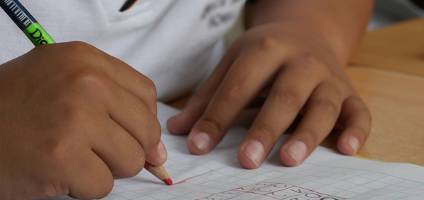A school pupil writing on a piece of paper with a red pencil.