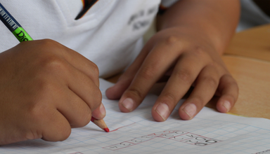 A school pupil writing on a piece of paper with a red pencil.