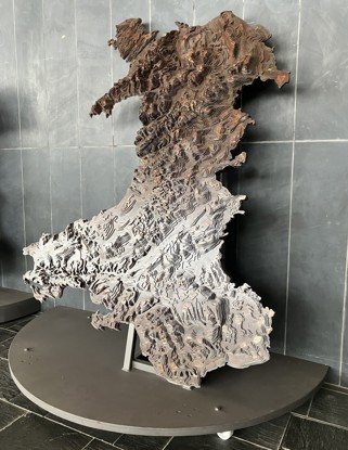 A steel relief map of Wales, showing the country's landscape through layered contour lines. The map is mounted on a metal stand.
