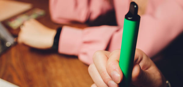 A person holding a green vape pen or e-cigarette. The focus is on the hand and the vape device 