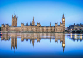 The UK Houses of Parliament in daytime from across the River Thames, with its reflection showing in the water. 
