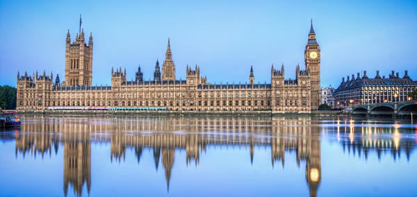 The UK Houses of Parliament in daytime from across the River Thames, with its reflection showing in the water. 