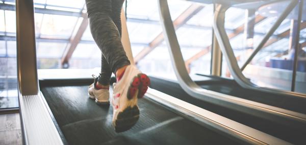 A person’s legs wearing sports trousers and trainers running on a treadmill with windows in the background.