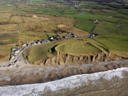 An image of the coastline taken from above. Waves create foam on the beach, some of the rocky cliff face is exposed and green fields are seen in the distance. 