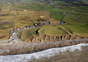 An image of the coastline taken from above. Waves create foam on the beach, some of the rocky cliff face is exposed and green fields are seen in the distance. 