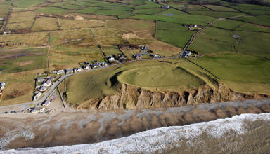 An image of the coastline taken from above. Waves create foam on the beach, some of the rocky cliff face is exposed and green fields are seen in the distance. 