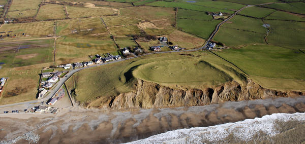 An image of the coastline taken from above. Waves create foam on the beach, some of the rocky cliff face is exposed and green fields are seen in the distance. 
