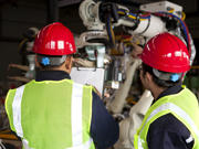 Two workers wearing hi-vis vests and hard hats looking at machinery while checking the contents of a document on a clipboard.