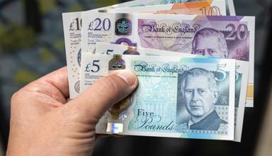 A hand holding a variety of UK bank notes, including £5, £10 and £20.
