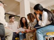 A group of people sitting in a circle in a brightly lit room, looking at papers and talking together during a collaborative discussion.