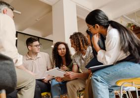 A group of people sitting in a circle in a brightly lit room, looking at papers and talking together during a collaborative discussion.
