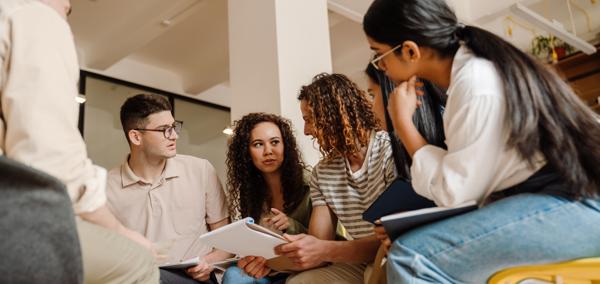 A group of people sitting in a circle in a brightly lit room, looking at papers and talking together during a collaborative discussion.