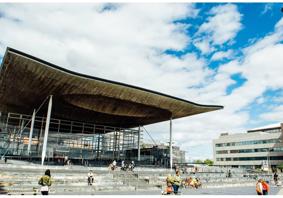 A photograph of the front of the Senedd building in Cardiff Bay. The building is comprised of grey bricks, and some white beams are visible. There is a sign that reads ‘Welsh Parliament’ in the back right hand side. In the foreground, there are pedestrians passing by. Some people are sitting on the steps outside the Senedd building.
