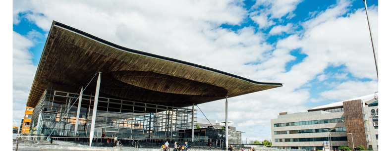 A photograph of the front of the Senedd building in Cardiff Bay. The building is comprised of grey bricks, and some white beams are visible. There is a sign that reads ‘Welsh Parliament’ in the back right hand side. In the foreground, there are pedestrians passing by. Some people are sitting on the steps outside the Senedd building.