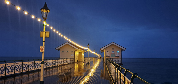 Glowing string lights reflect off a wet wooden pier at night, leading to two small buildings over the ocean