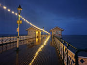 Glowing string lights reflect off a wet wooden pier at night, leading to two small buildings over the ocean