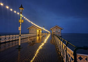 Glowing string lights reflect off a wet wooden pier at night, leading to two small buildings over the ocean