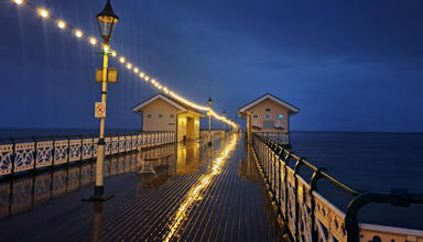 Glowing string lights reflect off a wet wooden pier at night, leading to two small buildings over the ocean