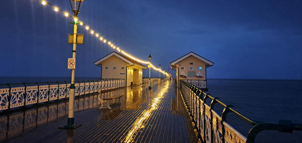 Glowing string lights reflect off a wet wooden pier at night, leading to two small buildings over the ocean