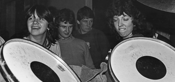 Black-and-white photograph of people gathered around large percussion instruments in a busy music venue.