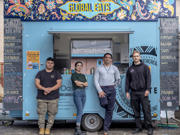 Four people stand in front of a turquoise colour food truck. The food truck has ‘Global Eats’ written large at the top and blackboard menus on the side. 