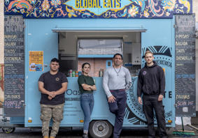 Four people stand in front of a turquoise colour food truck. The food truck has ‘Global Eats’ written large at the top and blackboard menus on the side. 