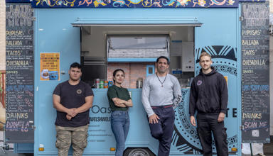 Four people stand in front of a turquoise colour food truck. The food truck has ‘Global Eats’ written large at the top and blackboard menus on the side. 