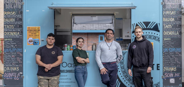 Four people stand in front of a turquoise colour food truck. The food truck has ‘Global Eats’ written large at the top and blackboard menus on the side. 