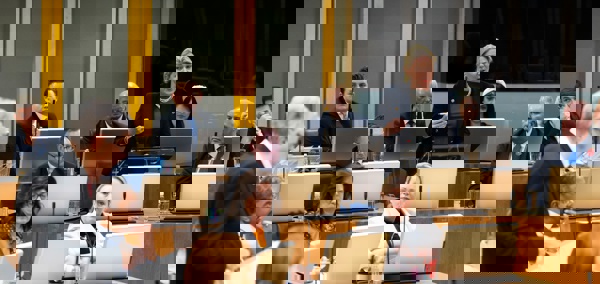 Mae Laura Ann Jones AS yn annerch y Prif Weinidog Mark Drakeford, sydd hefyd yn sefyll, yn ystod cyfarfod llawn. Mae aelodau eraill yn eistedd o'u cwmpas.