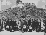 A grayscale image showing a large pile of rubble and debris in the background. In the foreground, a group of people are walking, carrying various bags and belongings.