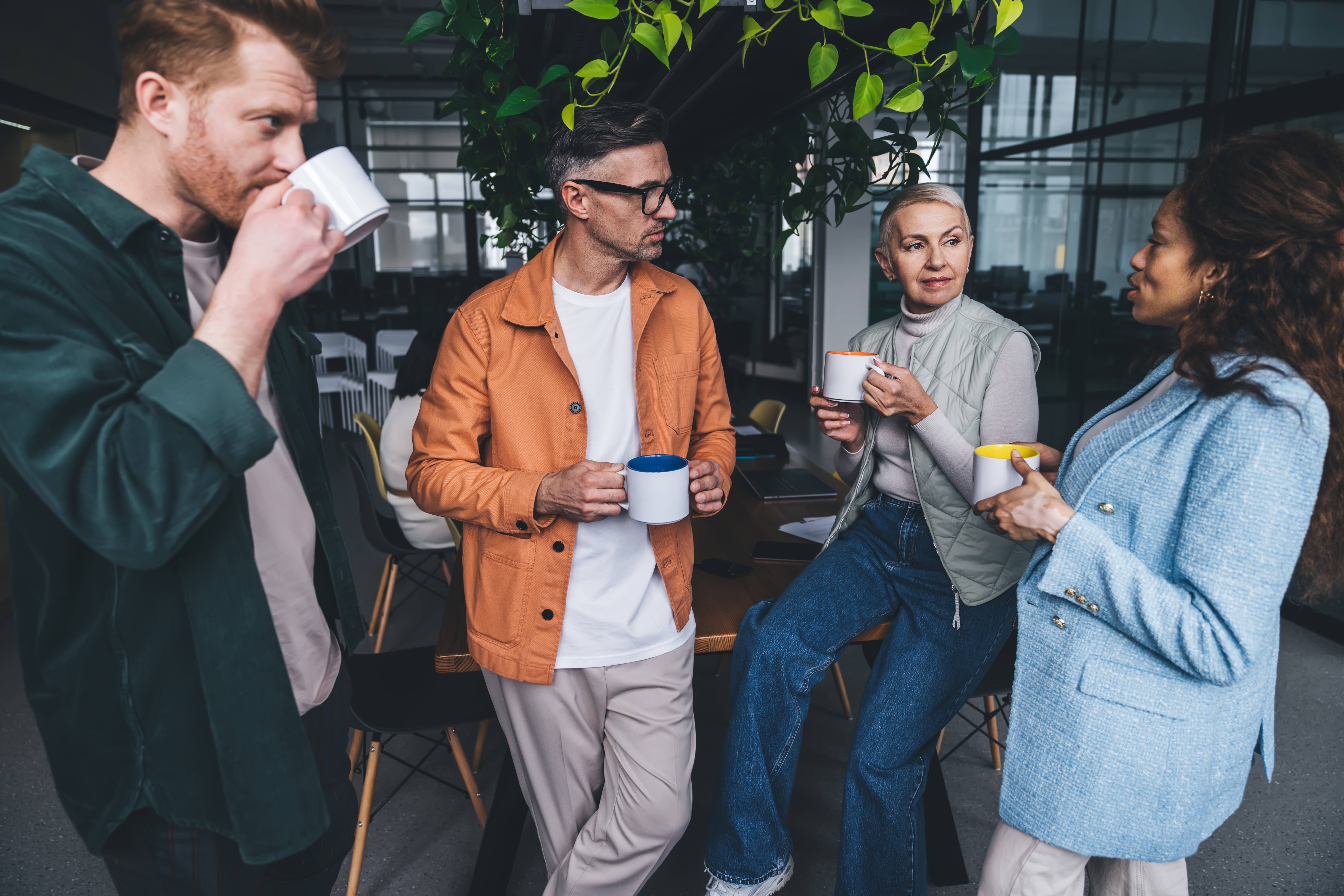 Four colleagues standing together in an office space, chatting and holding mugs.