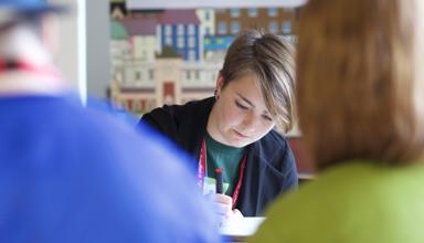 Image of a young woman writing whilst sat at a desk with other people blurred in the background
