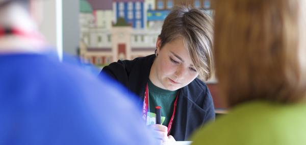 Image of a young woman writing whilst sat at a desk with other people blurred in the background