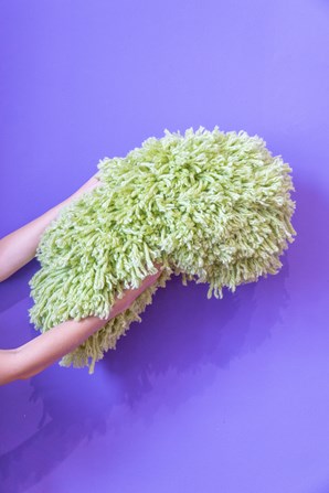 A person holding a fluffy green object in front of a bright purple background. The object is made of yarn and is round and long in shape.