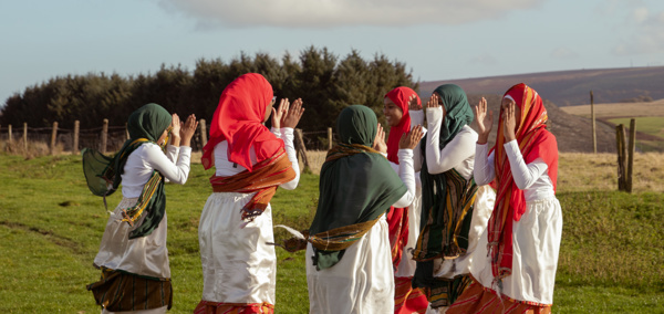 A group of young women wearing traditional clothing and headscarves, standing in a circle dancing on a grassy field with trees, hills, and a partly cloudy sky in the background.